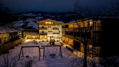 Beleuchtetes Hotel mit verschneitem Spielplatz und Schaukeln vor einer Berglandschaft bei Nacht