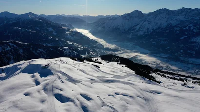 Verschneite Skipisten mit Blick auf ein breites Alpental und schneebedeckte Berge